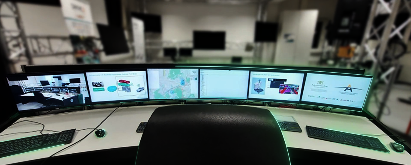 Vor einem leeren Bürostuhl sind mehrere PC-Monitore auf einem Schreibtisch aufgestellt, die verschiedene Themen rund um autonomes Fahren abbilden.