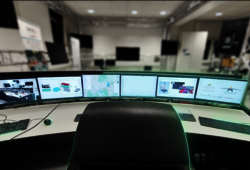 Vor einem leeren Bürostuhl sind mehrere PC-Monitore auf einem Schreibtisch aufgestellt, die verschiedene Themen rund um autonomes Fahren abbilden.