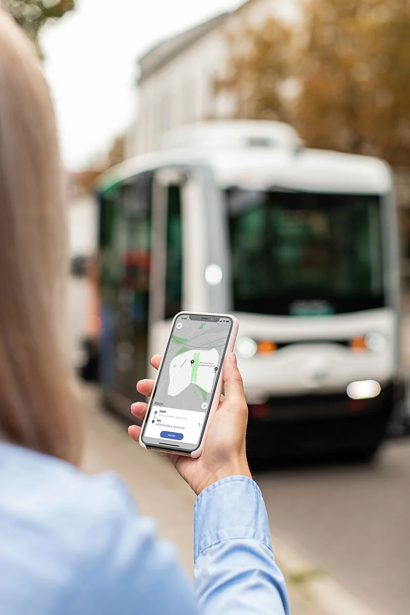 Eine Frau hält ein Smartphone mit geöffneter Mobilitäts-App, im Hintergrund steht ein autonomer Shuttlebus.
