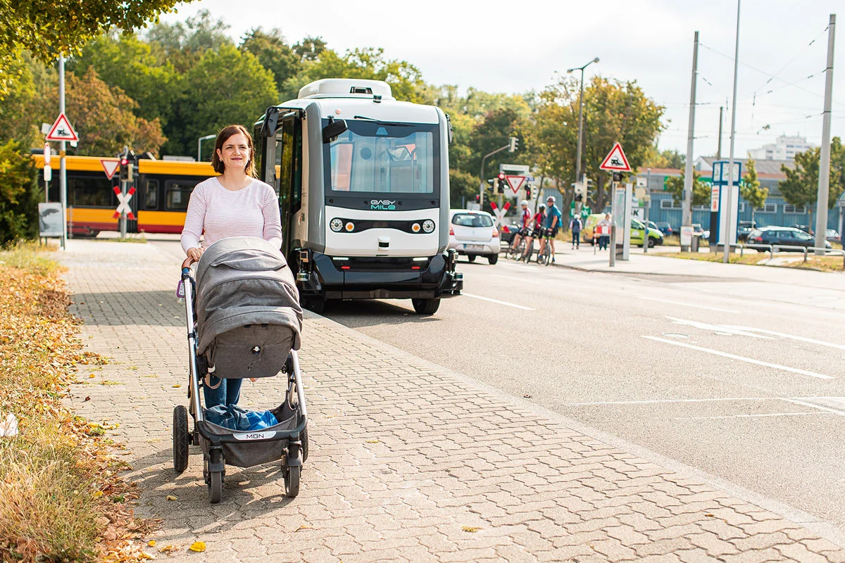 Eine Frau schiebt einen Kinderwagen auf dem Gehweg, im Hintergrund fährt ein autonomer Shuttlebus.